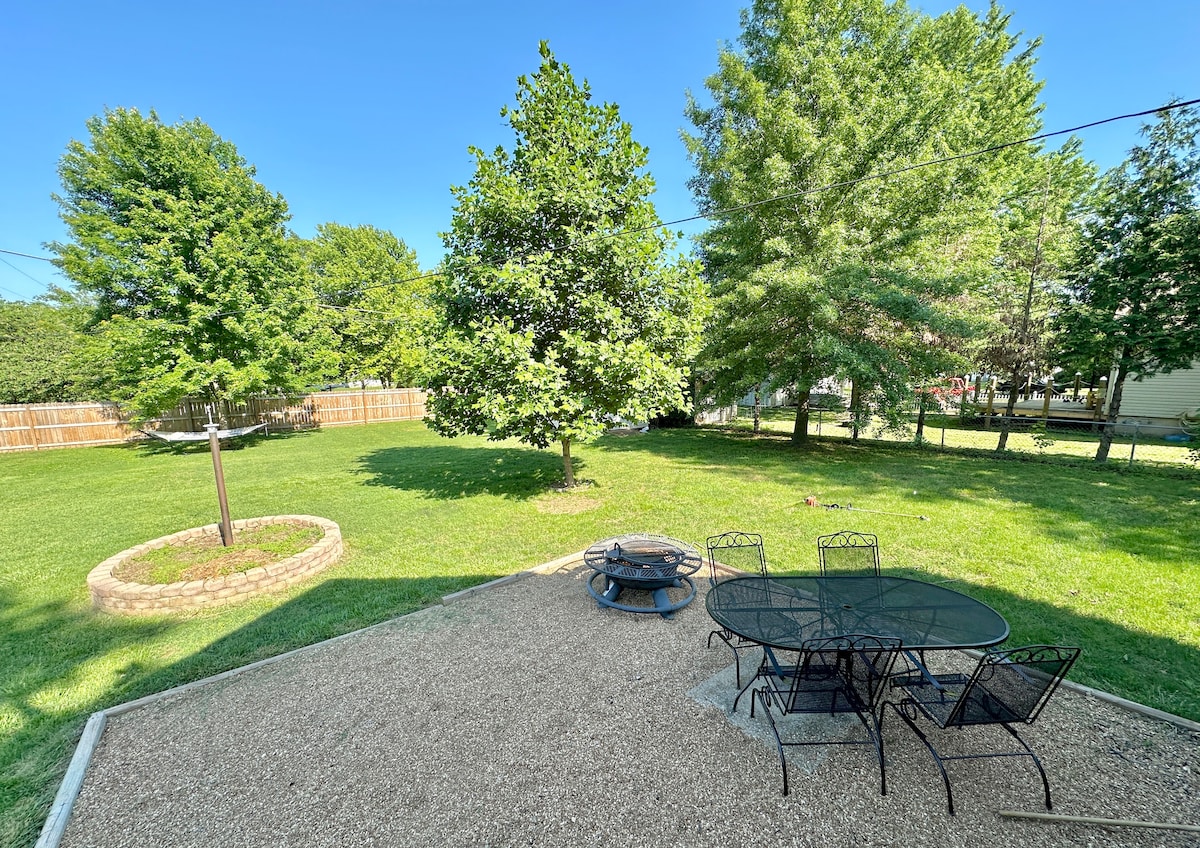 A spacious backyard is displayed, featuring a gravel area with a round fire pit and a metal dining set with four chairs. Lush green grass surrounds the setting, complemented by mature trees providing shade. A wooden fence marks the perimeter of the yard.