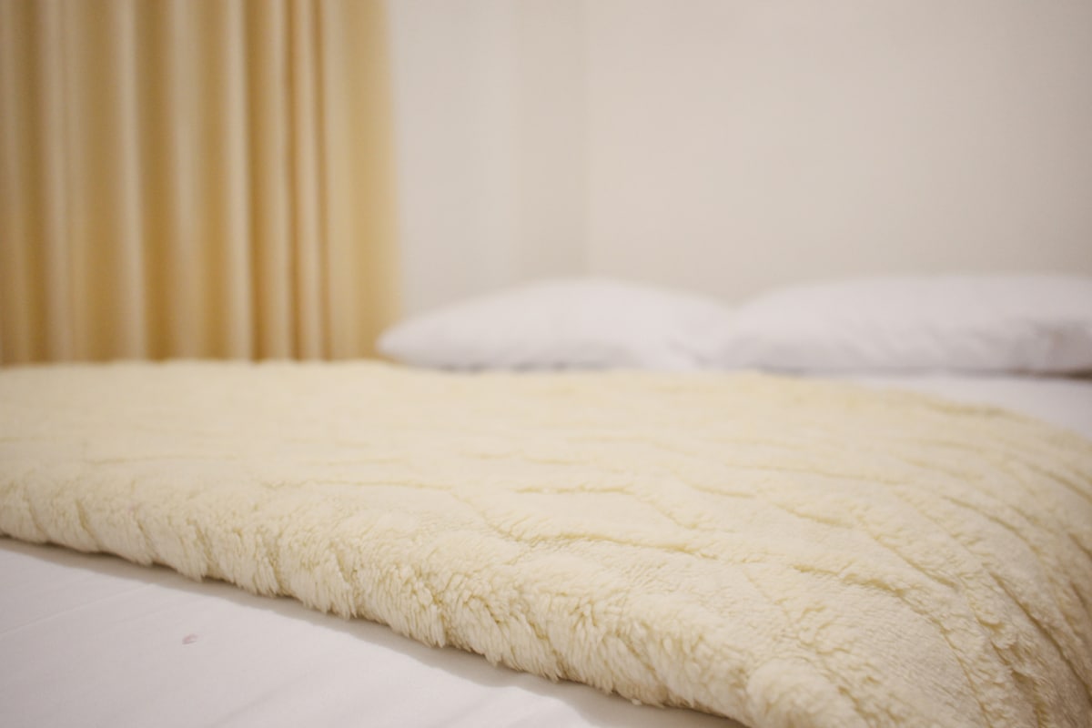 A soft, textured yellow blanket is placed over a spacious bed covered with clean white linens. Pillows are neatly arranged at the head of the bed, with light-colored curtains softly draping in the background.
