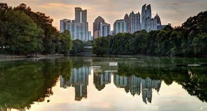 A view of a city skyline reflecting on a tranquil lake is presented. Skyscrapers are visible, surrounded by lush greenery, creating a contrast between urban structures and natural elements. The sky displays soft hues of orange and blue as daylight transitions to evening.