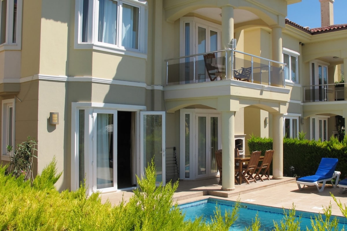 A three-story villa showcases a private swimming pool surrounded by greenery. The exterior features large glass doors leading to outdoor seating and a balcony on the upper level. Sun loungers are positioned near the pool area, enhancing the tranquil outdoor space.
