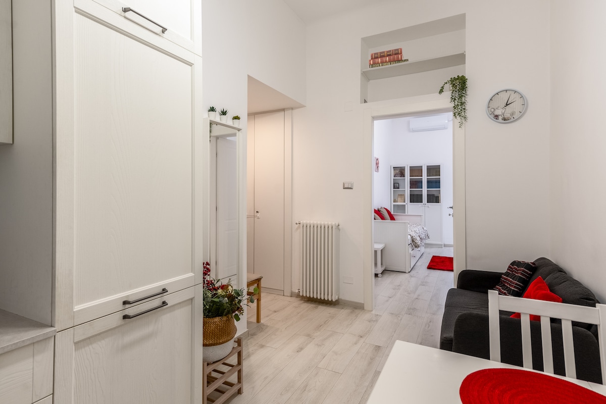 A bright and airy living space is captured, featuring light-colored flooring and white walls. The room showcases a cozy black sofa against the wall, with decorative elements like a small plant and a wall clock adding warmth to the ambiance. Access to other rooms is visible through the doorway.