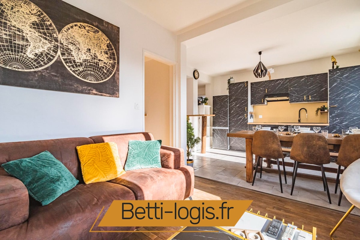 A inviting living area features a brown sofa adorned with colorful cushions. A dining table set for meals is visible in the background, surrounded by brown upholstered chairs. The space is illuminated by natural light, highlighting a modern kitchen with a sleek design.