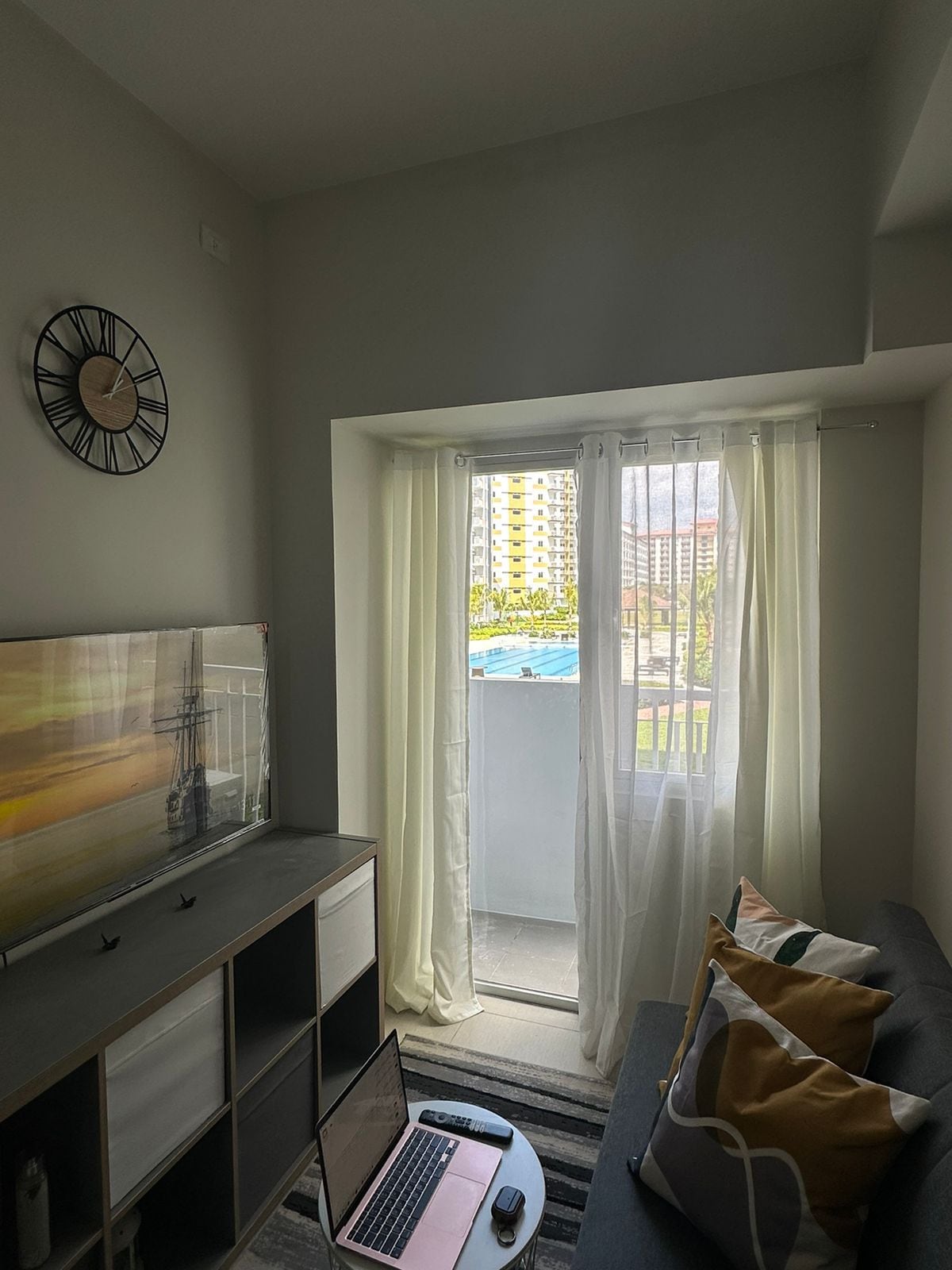 A compact living area features a gray sofa adorned with decorative cushions, complemented by a sleek television mounted on the wall. Natural light floods the space through sheer white curtains, with a glimpse of the pool view visible through the balcony door.