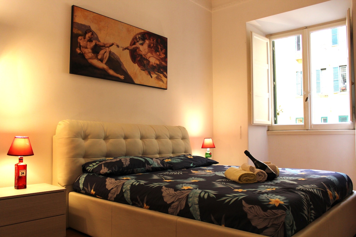 A spacious bedroom displays a queen-sized bed adorned with a floral-patterned bedspread. Soft lighting is provided by bedside lamps, and a large artwork hangs above the bed. Natural light enters through a wide window, enhancing the room's brightness.