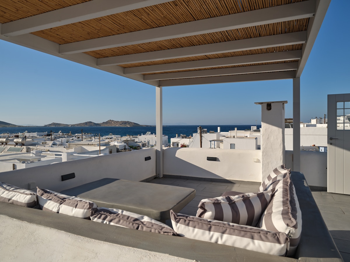 An open terrace area is highlighted, featuring a comfortable seating arrangement with cushioned benches and soft pillows. The space is shaded by a wooden overhead structure, providing a picturesque view of the sea and surrounding whitewashed buildings typical of the Cycladic architecture.