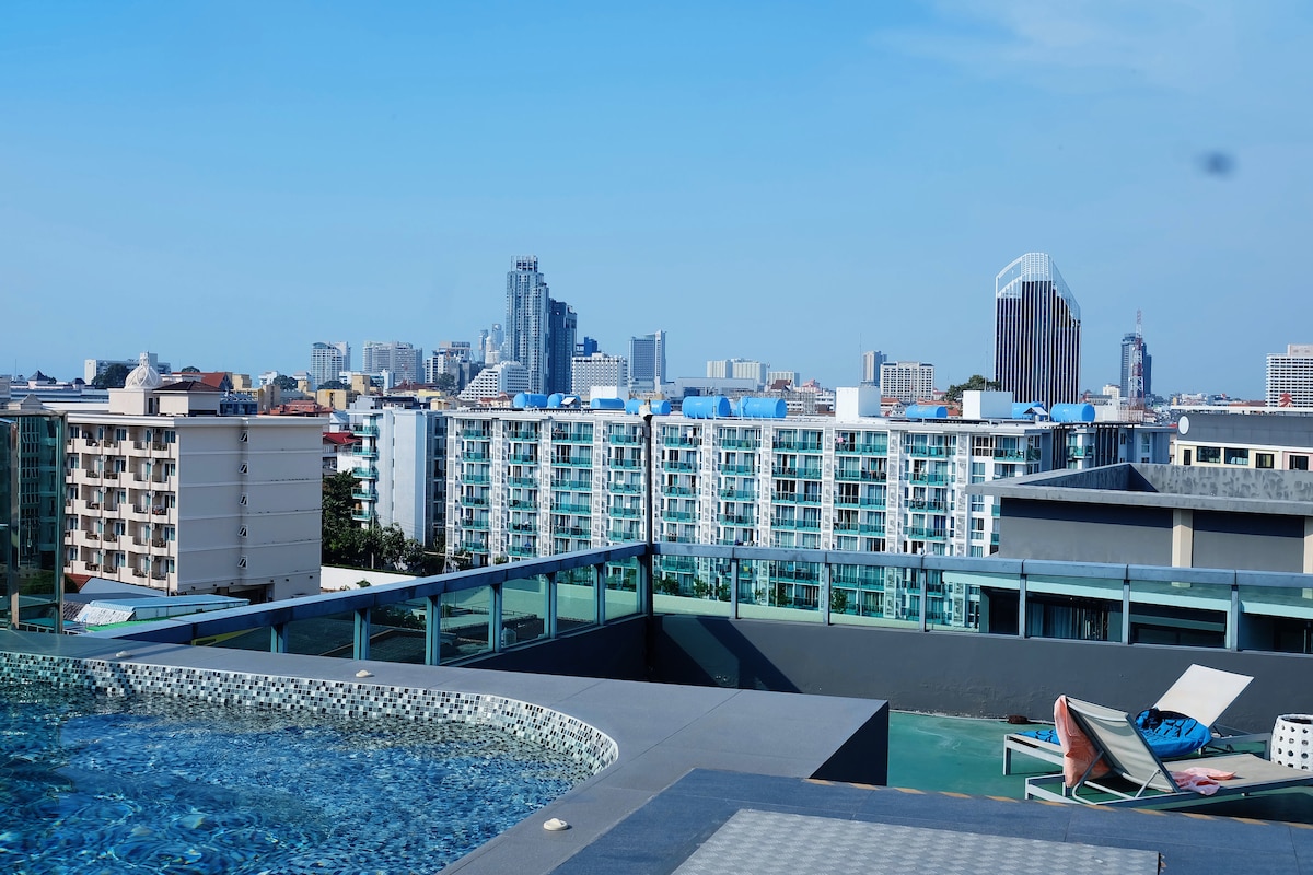 A rooftop area is featured, showcasing a swimming pool with a city skyline in the background. The view includes various modern buildings and structures, with seating options available near the pool and a clear sky above.