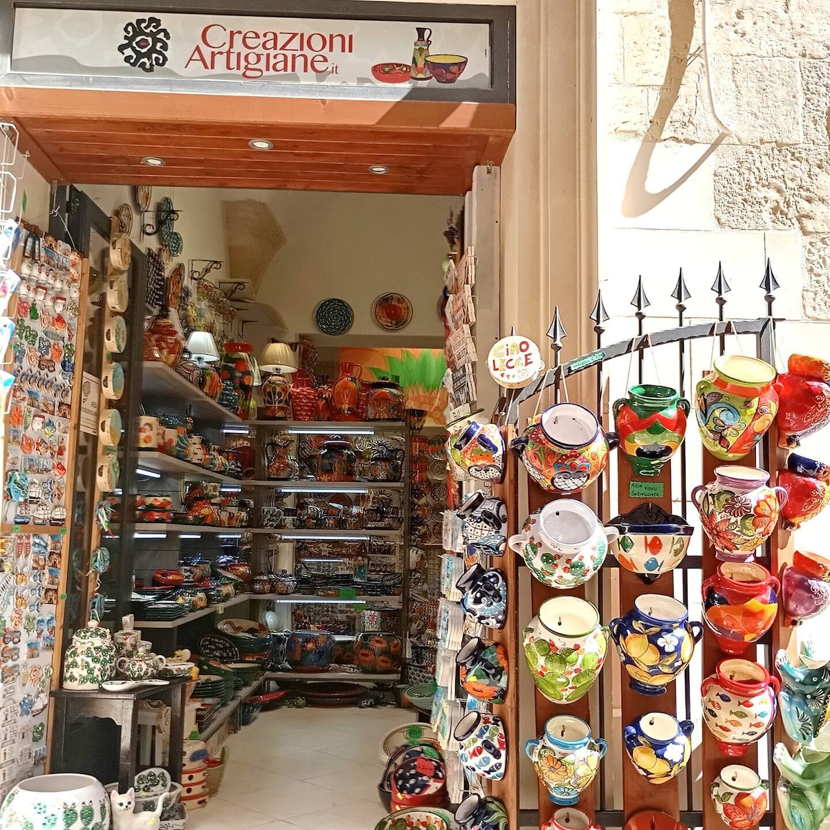 The entrance of a colorful artisan shop is displayed, framed by a wooden sign. Vibrant ceramic items, including plates and mugs, are neatly arranged on shelves inside. A wall-mounted display showcases hanging pottery, adding to the inviting atmosphere of local craftsmanship.