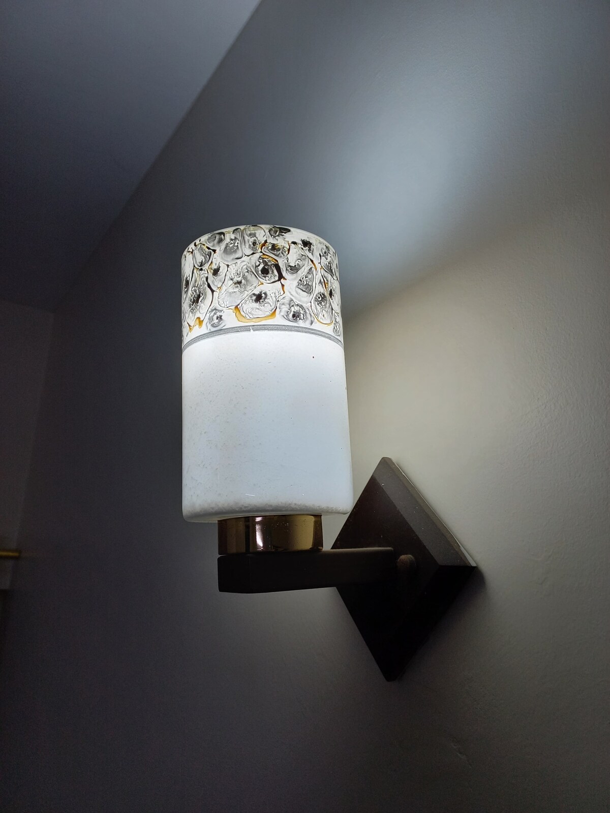 A wall-mounted light fixture is displayed, featuring a cylindrical glass shade with a metallic decorative band at the top. The light emits a soft glow, creating a gentle illumination on the adjacent wall.