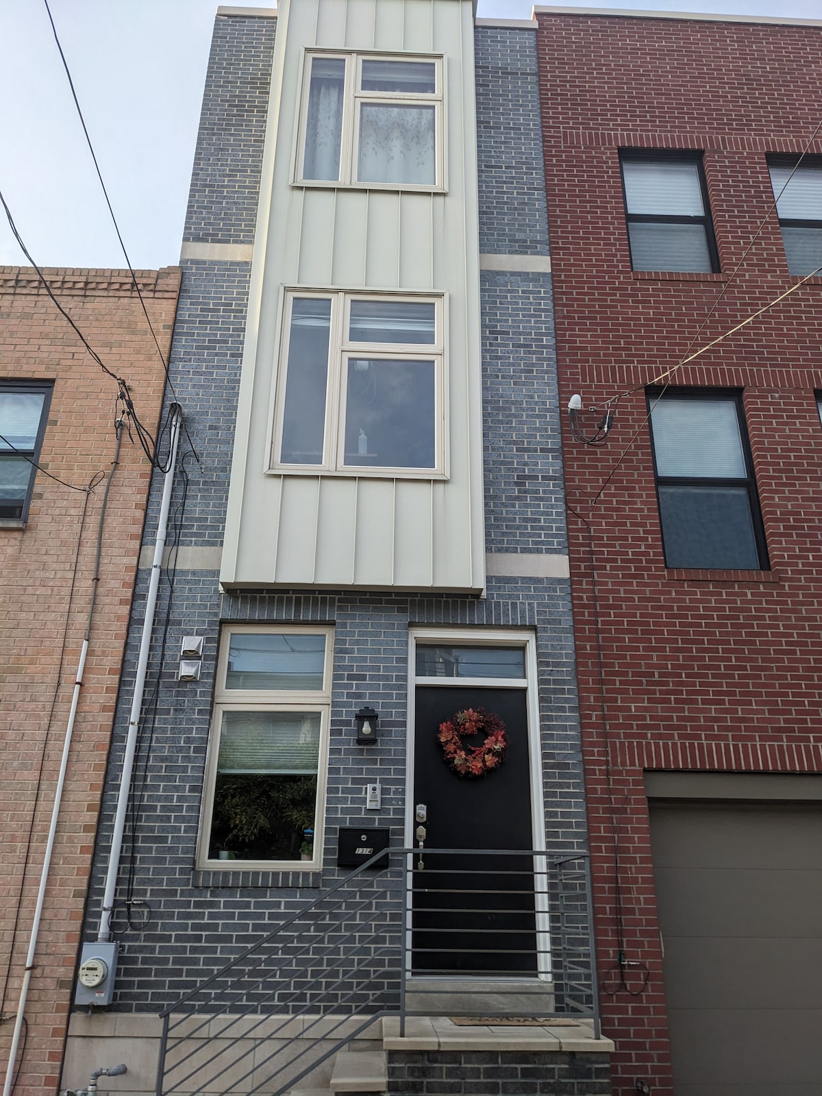 The exterior of a three-story house is shown, featuring a combination of brick and gray paneling. A modern black front door is framed with a decorative wreath, and large windows provide natural light. A small set of stairs leads to the entrance, which is accentuated by a railing.
