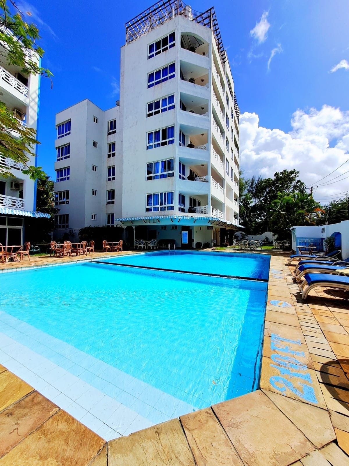 A bright pool area features a large, clear swimming pool surrounded by lounge chairs. The modern building stands nearby, with large windows reflecting the sunlight. The setting is complemented by green foliage, adding to the inviting atmosphere.