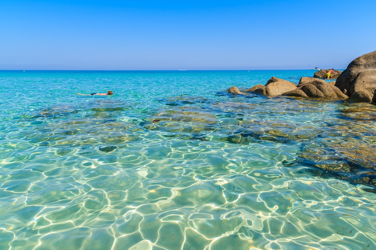 A calm sea is captured, showcasing clear turquoise waters that gently lap against a rocky shore. Sunlight reflects off the water's surface, creating a shimmering effect. The horizon meets a clear blue sky, providing a tranquil coastal scene.