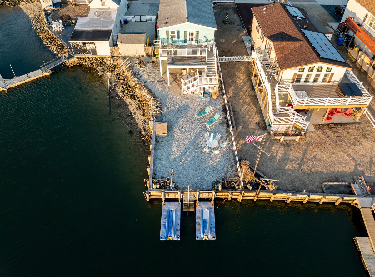 An aerial view captures the Bay Breeze Retreat, situated along a serene waterway. The property features a large, gravel patio with lounge chairs overlooking the water and a private dock with two boat slips. Nearby homes and outdoor spaces reflect a relaxed coastal vibe.