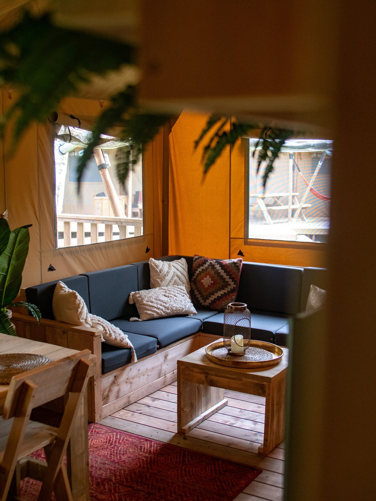 A comfortable seating area is arranged with a dark sofa adorned with patterned cushions. A rustic wooden coffee table sits at the center, accompanied by a candle. Natural light filters through two windows, illuminating the warm tones of the wooden floor.