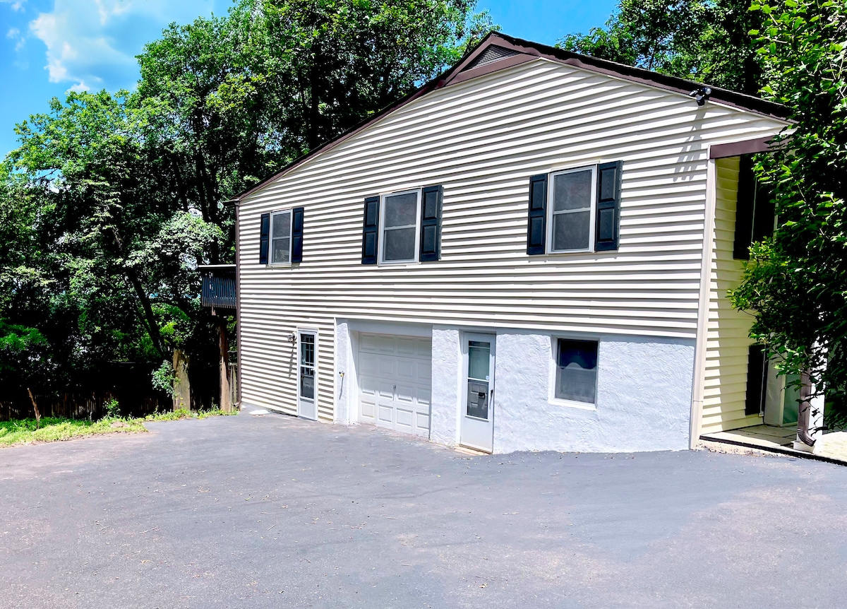 A spacious two-story house is presented, characterized by a light-colored exterior with dark shutters. A large driveway leads to a garage door on the left, while a pathway is visible on the right, surrounded by green foliage.