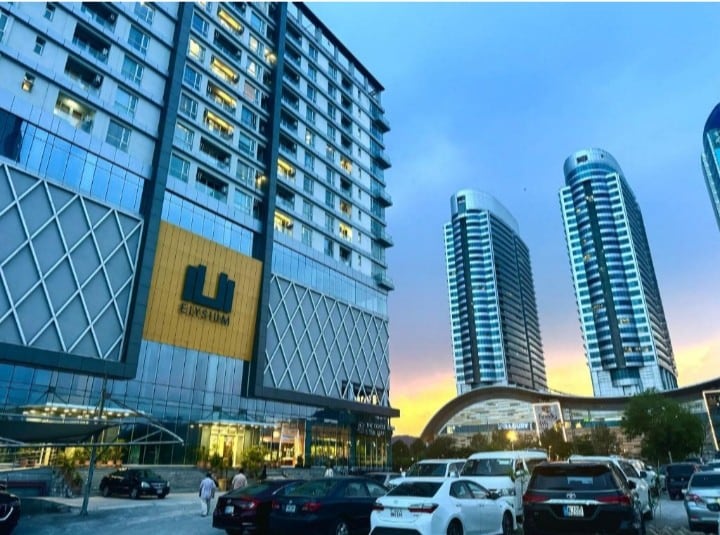 An urban landscape showcases modern high-rise buildings with reflective glass façades. Several vehicles are parked in a spacious lot, with a bright sunset casting a warm glow in the background. Architectural features blend contemporary design with functional spaces.
