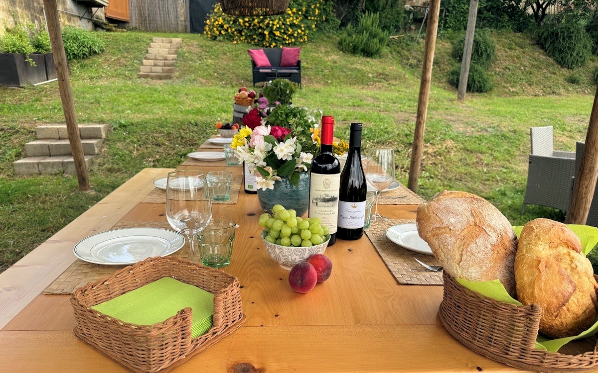 A spacious outdoor dining table is set under a pergola, displaying an array of glassware, plates, and woven baskets filled with bread and fruit. Fresh flowers are featured in a central bowl, while a scenic garden is visible in the background, complemented by comfortable seating.