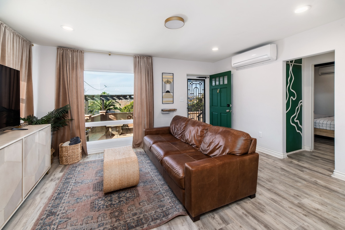 A spacious living area is highlighted by a brown leather sofa and an area rug. Natural light filters through large windows with elegant curtains, revealing a view of the patio. A flat-screen TV is mounted above a sleek entertainment unit, contributing to the modern design.