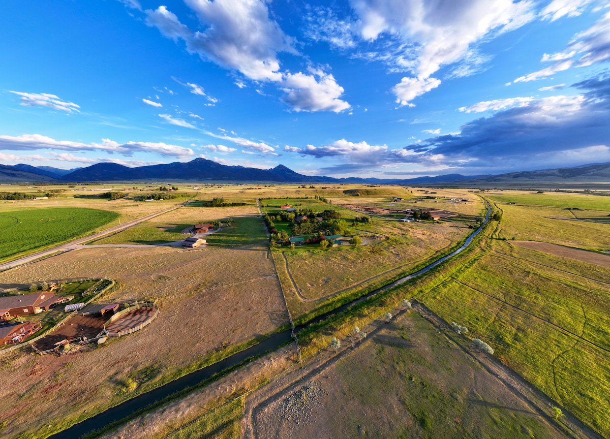 An expansive aerial view of a tranquil landscape showcases rolling hills and fields under a vast sky filled with clouds. The scenery includes distant mountains and a meandering river, leading through the golden and green fields that characterize the serene setting of Paradise Valley.