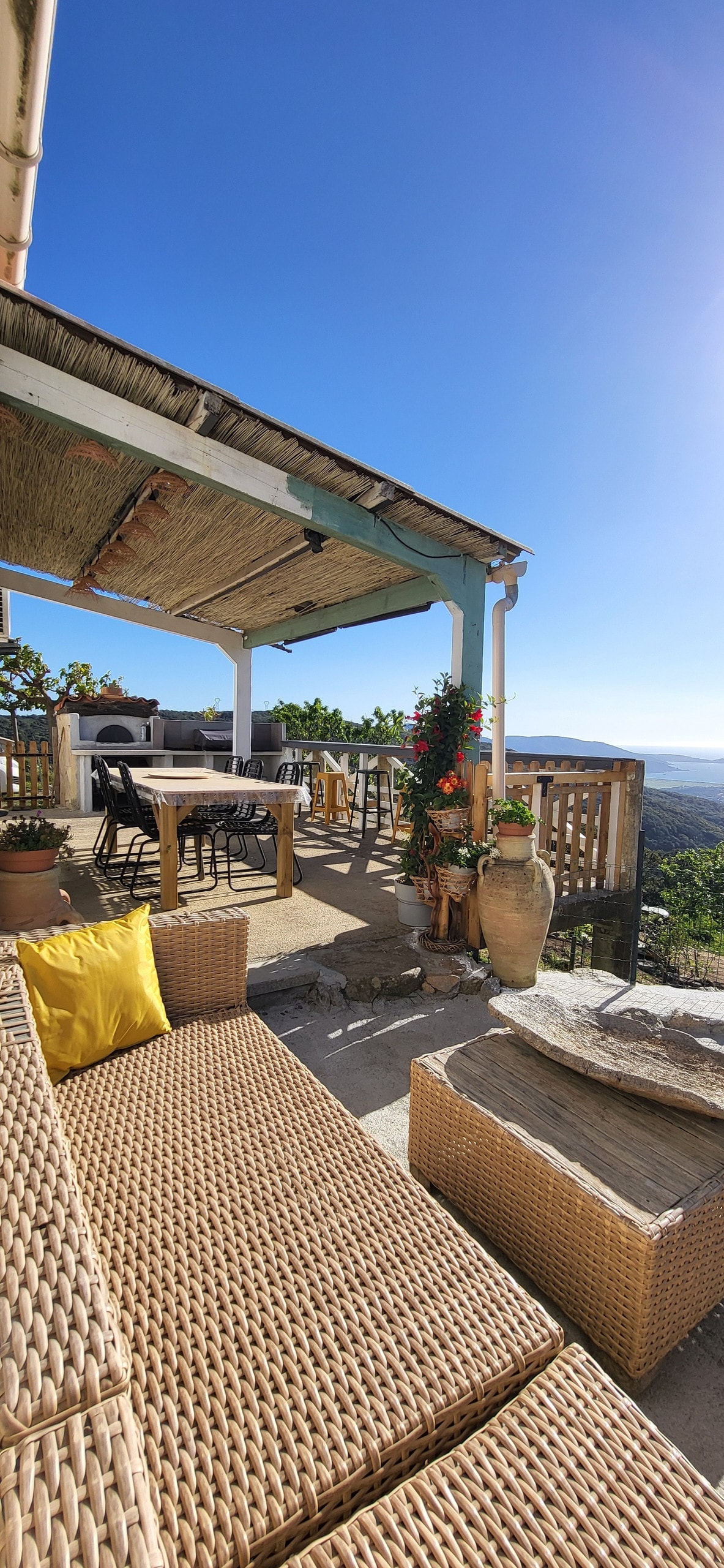 A spacious terrace is highlighted, featuring a dining area and comfortable seating. Lush greenery and decorative plants surround the space, enhancing the view of the landscape. A decorative vase adds a charming touch, and blue skies are visible above.