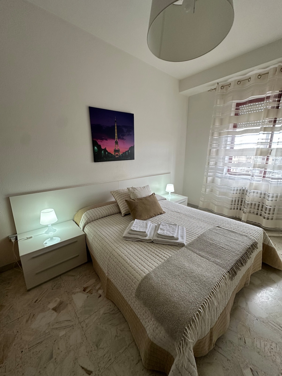 A cozy bedroom is presented with a spacious bed covered in a textured blanket, illuminated by two bedside lamps. A piece of wall art depicting a sunset is displayed above the bed. Curtains filter natural light from the window, enhancing the room's soft ambiance.
