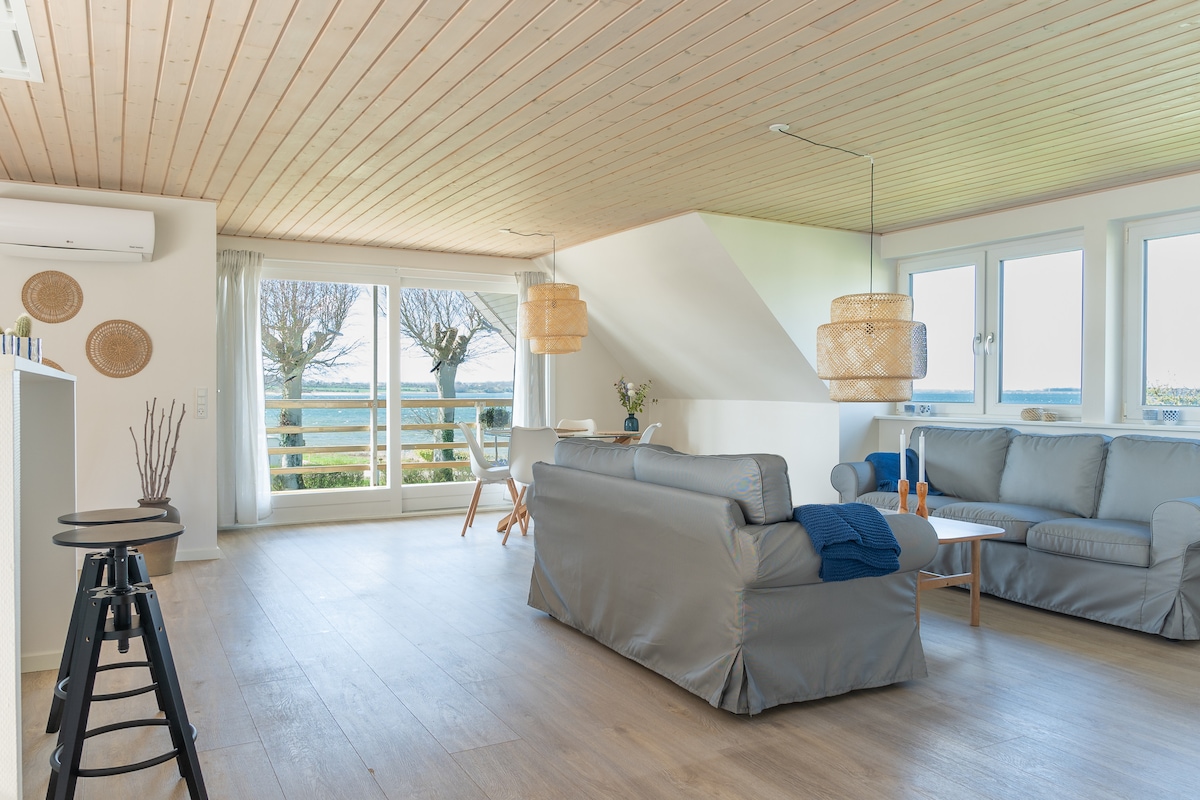 A bright living area features large windows that provide views of the water. Soft furnishings in neutral tones are complemented by wooden ceiling beams. Two stylish pendant lights hang above a cozy seating arrangement, and a small table is set nearby, enhancing the inviting atmosphere.