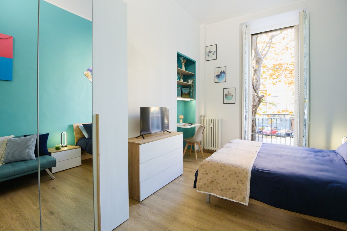 An open space is depicted with a spacious bed covered in a dark blanket. A sleek dresser and a Smart TV are present, while a comfortable seating area features a sofa. Natural light enters through a nearby window, highlighting the bright blue wall.