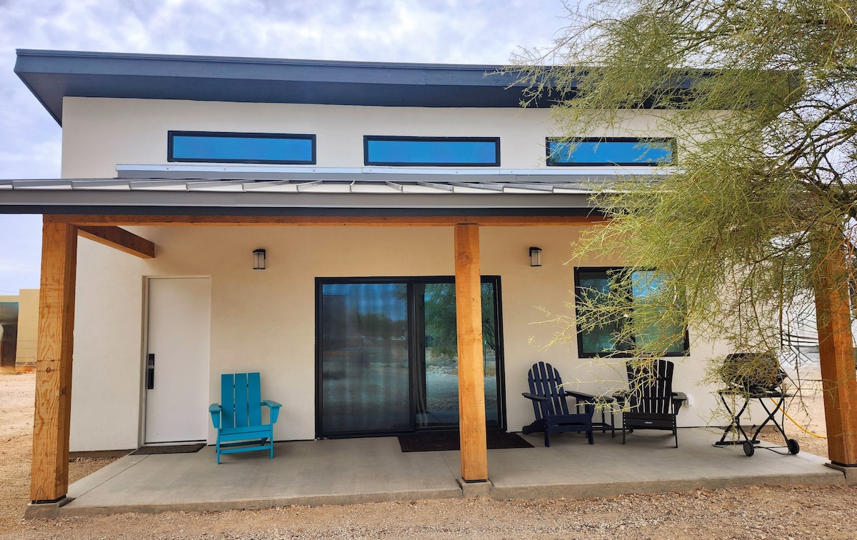 A modern guest house exterior is showcased with a covered porch featuring two light blue chairs and a small bistro table with matching chairs. Large windows allow natural light to enter, while a saguaro cactus is visible nearby, set against a sandy landscape.