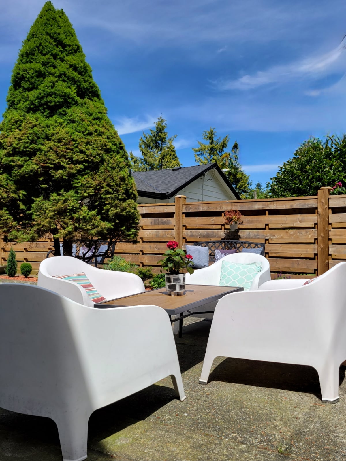 A private outdoor courtyard area is highlighted by a small table surrounded by four white chairs. Lush greenery and a tall tree create a serene backdrop, complemented by wooden fencing, while bright flowers add a touch of color to the space.
