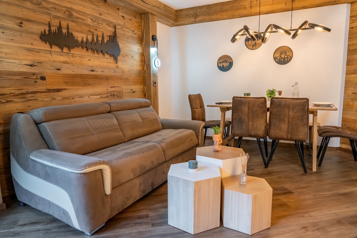 A spacious living area features a comfortable sofa in soft brown tones, accompanied by two geometric wood tables. A dining table with dark upholstered chairs is positioned nearby, while a unique overhead light fixture adds a modern touch. The wooden wall enhances the natural ambiance.