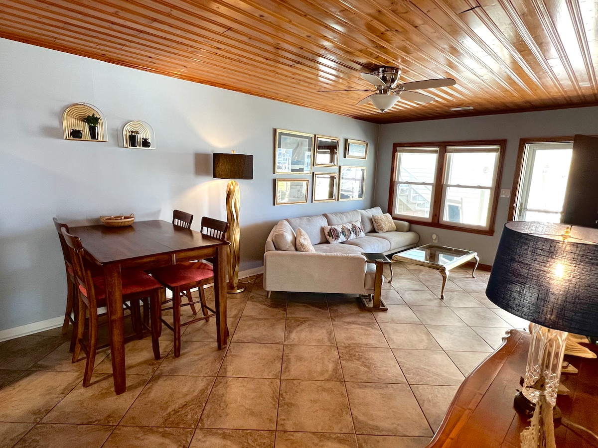 An inviting living area features a cozy couch accompanied by decorative accent pillows. A wooden dining table with four chairs provides a gathering space. Natural light flows in from multiple windows, highlighting the warm wooden ceiling and neutral wall colors, creating a comfortable atmosphere.