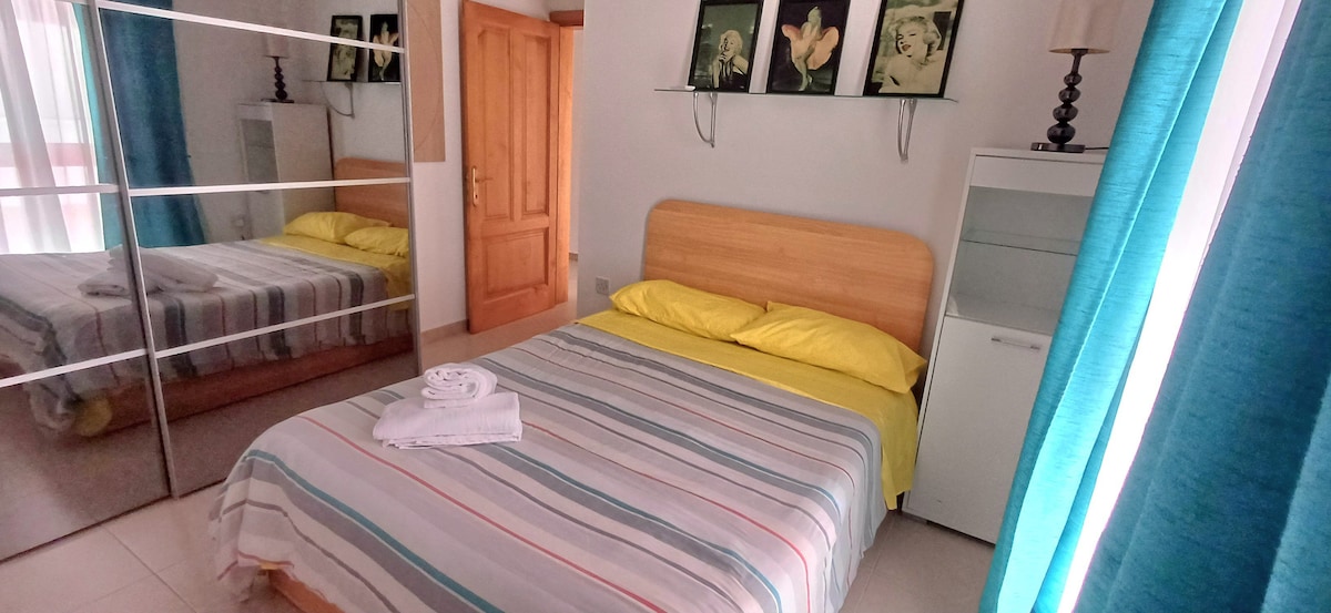 A sleeping area features a wooden bed frame with a striped blanket and bright yellow pillows. A mirror-fronted wardrobe reflects the space, while a white unit and a lamp are positioned nearby. Light filters through sheer curtains hung by a window.