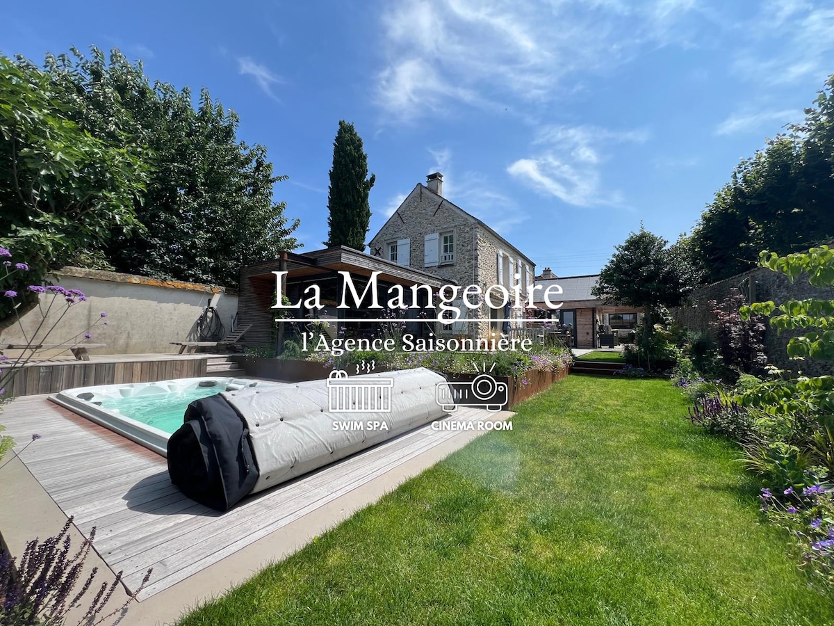 The outdoor area features a landscaped garden with a well-maintained lawn, bordered by greenery. A swim spa is visible on the terrace, adjacent to the charming stone house. The sky is clear, suggesting a bright and inviting atmosphere.