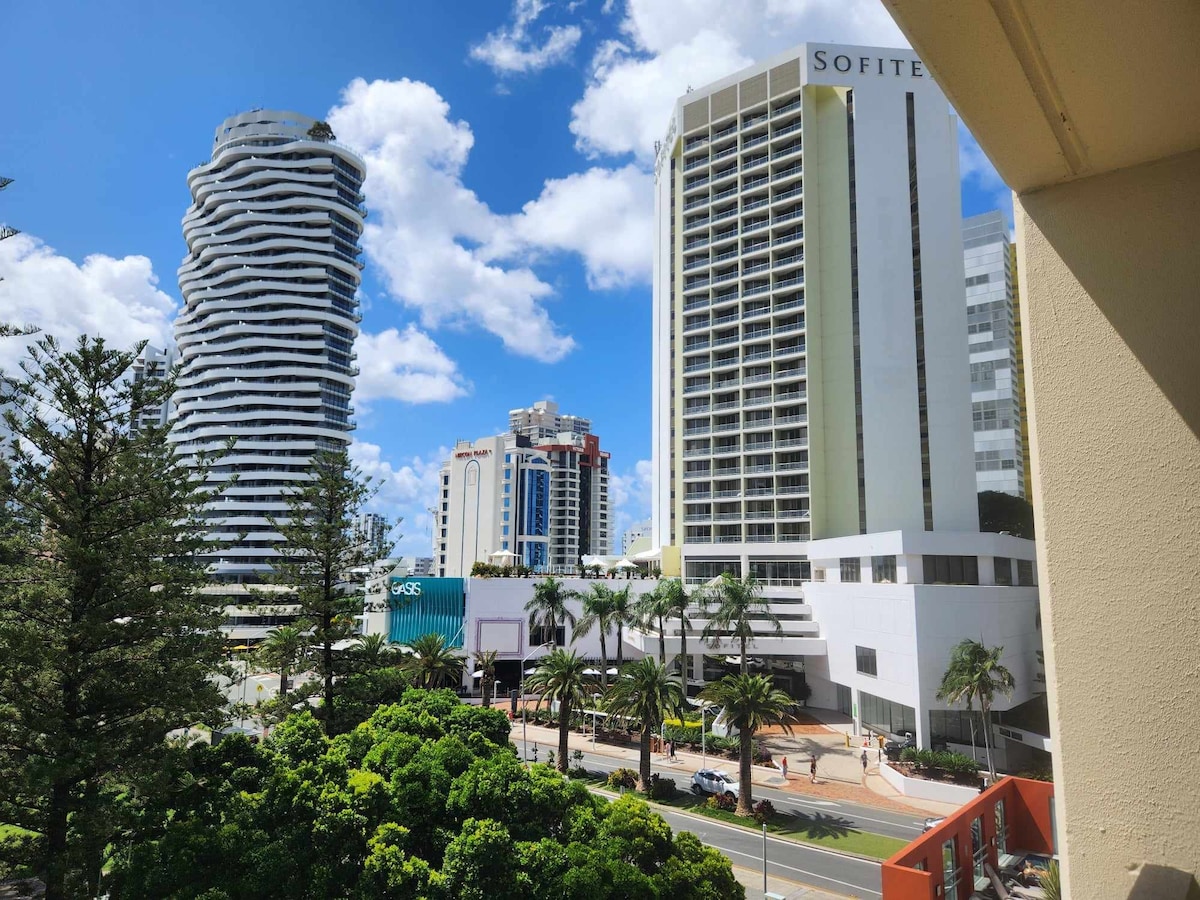 A vibrant view showcases modern high-rise buildings against a bright blue sky. Lush greenery is visible in the foreground, providing contrast to the urban landscape. Nearby palm trees line the streets, enhancing the lively atmosphere of the Broadbeach area.