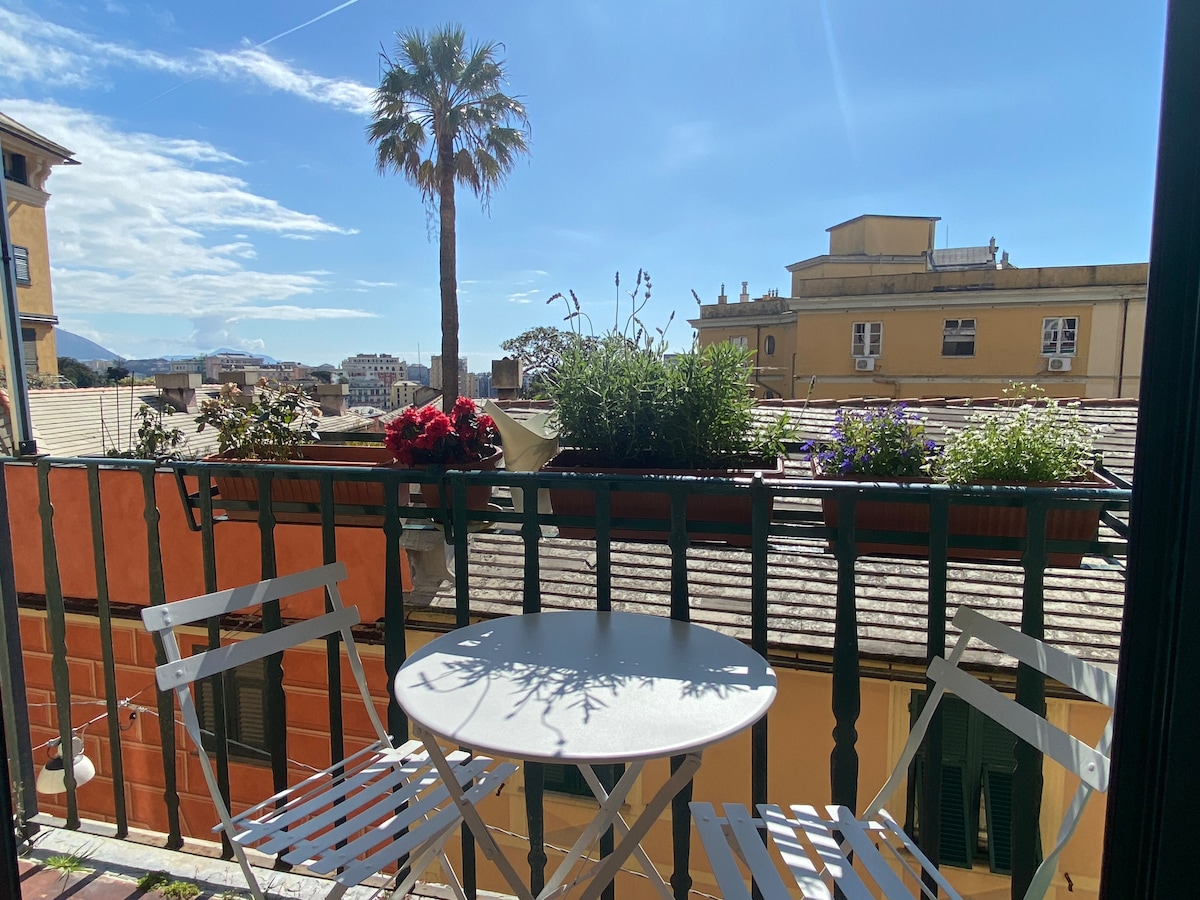 A small balcony is visible, featuring two white metal chairs and a round table. Potted plants with vibrant flowers line the railing. The view includes rooftops and distant hills against a clear blue sky, providing a serene outdoor space.