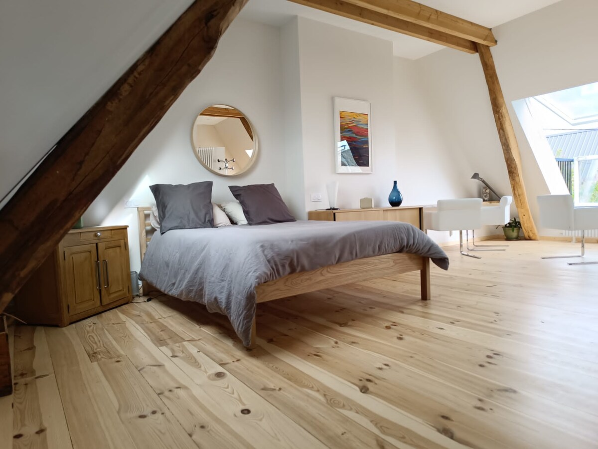 A spacious bedroom is featured, with a wooden bed dressed in grey linens. Two large windows and a circular mirror reflect light, enhancing the airy feel. Natural wood beams and flooring add warmth, complemented by simple, modern furniture including a small side table and two chairs.