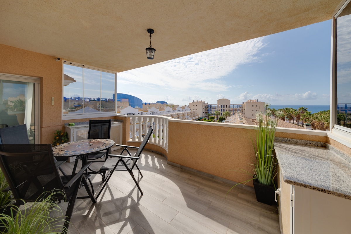 A spacious terrace features a round table surrounded by four black chairs, with vibrant greenery in planters adding a natural touch. An unobstructed view showcases the surrounding buildings and distant sea, while large windows allow natural light to fill the area.