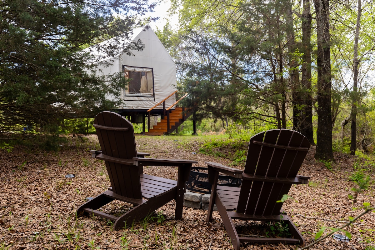 Two wooden adirondack chairs are positioned around a fire pit, surrounded by a natural landscape of trees and fallen leaves. A glamping tent is visible in the background, elevated on a wooden platform, with stairs leading up to its entrance.