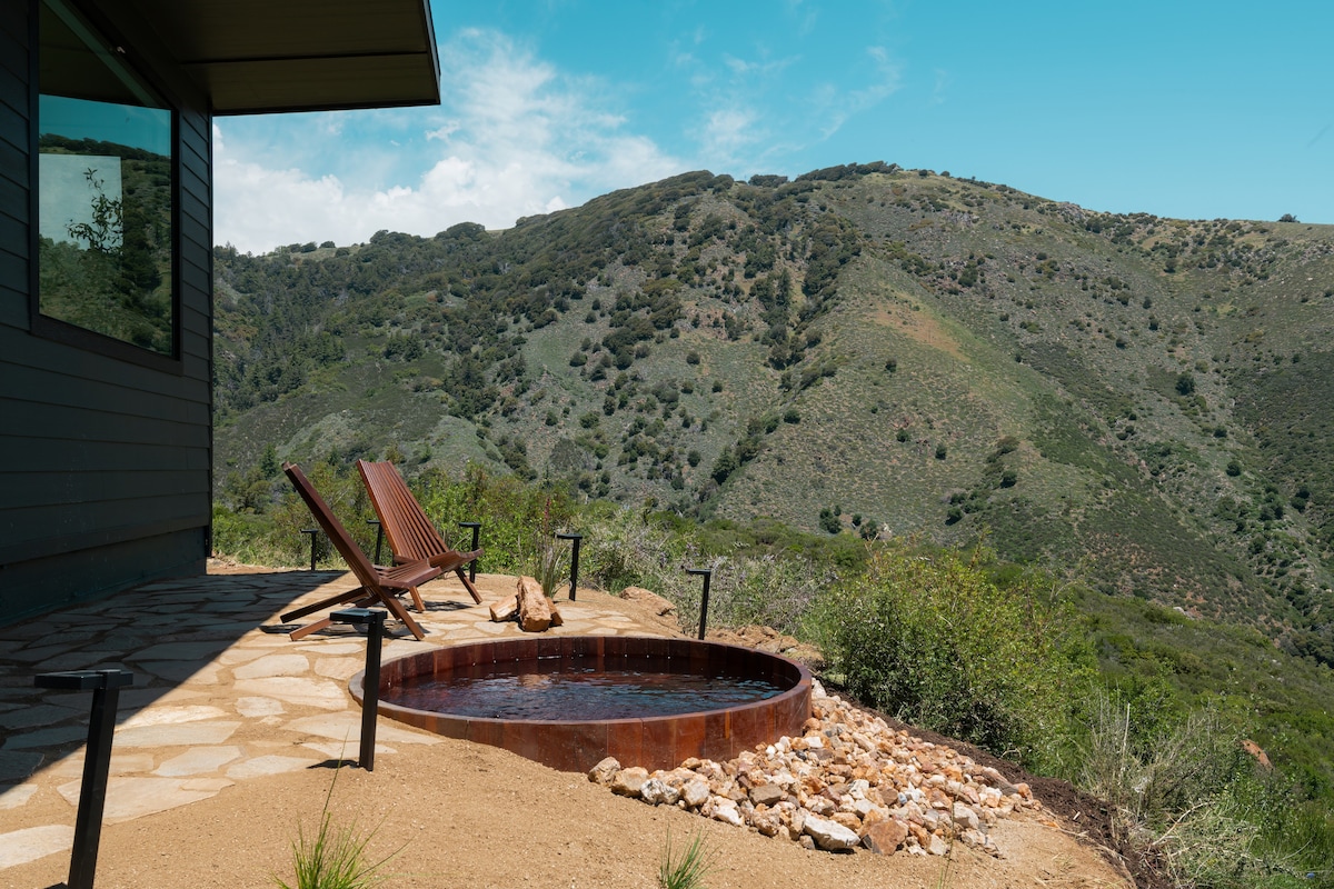 A custom cedar soaking tub is nestled in a rocky area, surrounded by natural landscaping. Two wooden lounge chairs are positioned nearby, offering views of the rolling hills and greenery in the distance. A clear blue sky completes the serene outdoor setting.