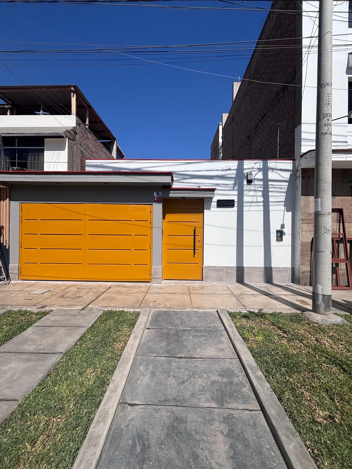 A modern exterior view of the apartment is displayed, featuring a bright yellow garage door and entrance. The building is framed by clear blue skies and is surrounded by a neatly paved pathway and green grass.