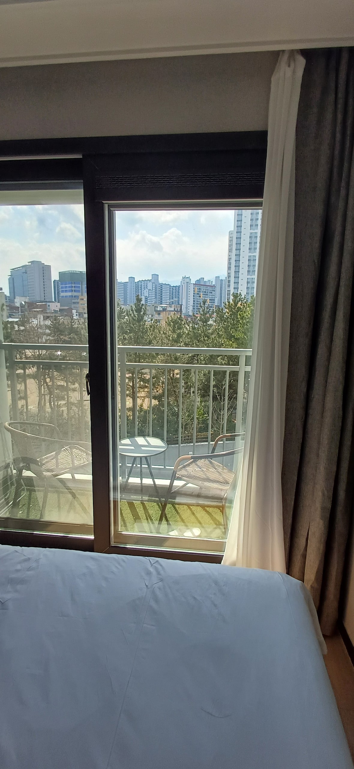 A spacious room features large glass doors leading to a balcony, offering city views. Two outdoor chairs are visible on the balcony, with greenery seen in the foreground. Soft natural light filters in through the curtains, illuminating the comfortable bedding.