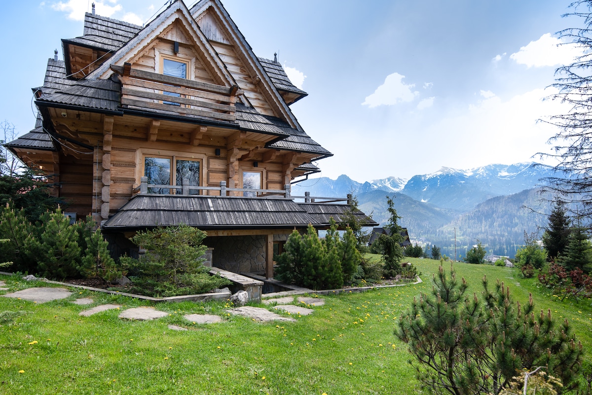 A charming wooden house features a sloped roof and a spacious balcony, surrounded by small hedges and greenery. The backdrop showcases expansive mountain views under a partly cloudy sky, creating a serene outdoor setting.