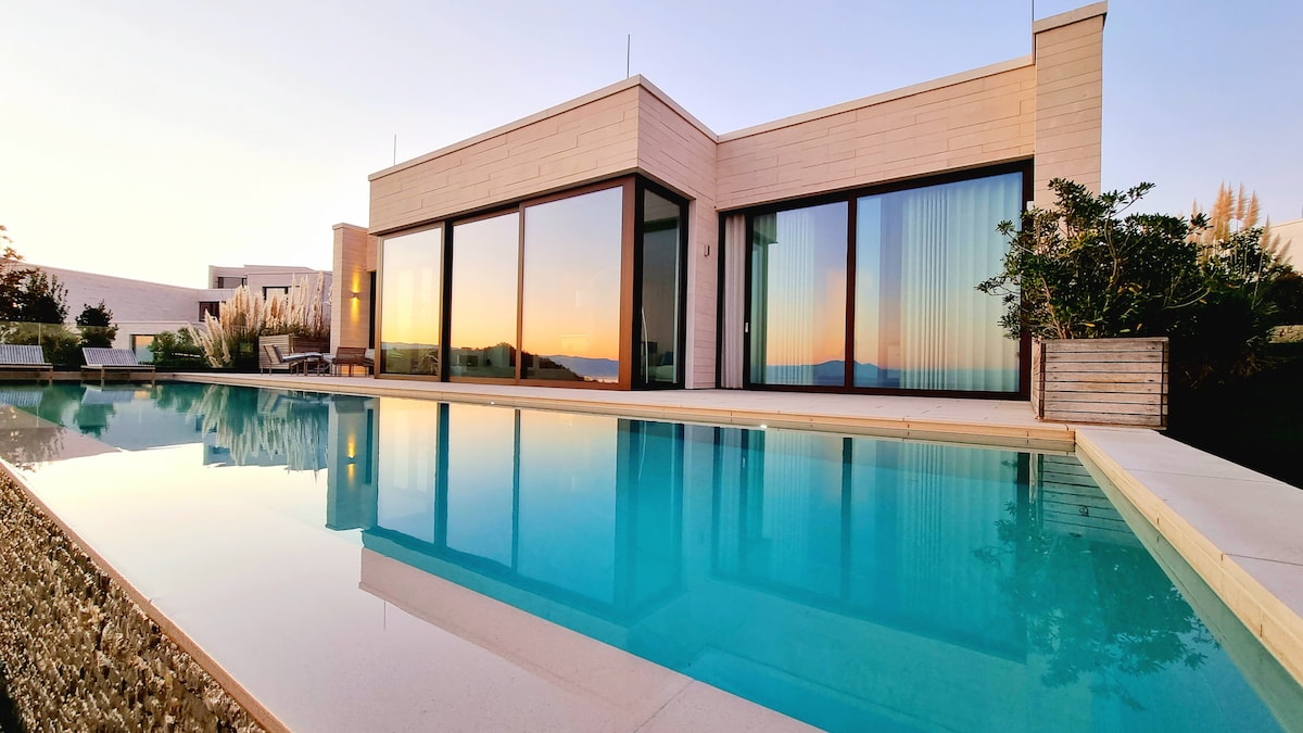 The villa's exterior is showcased with large glass windows reflecting the serene sunset. An infinity pool flows seamlessly alongside the structure, mirroring the tranquil water. Lush greenery surrounds the area, enhancing the connection with nature.