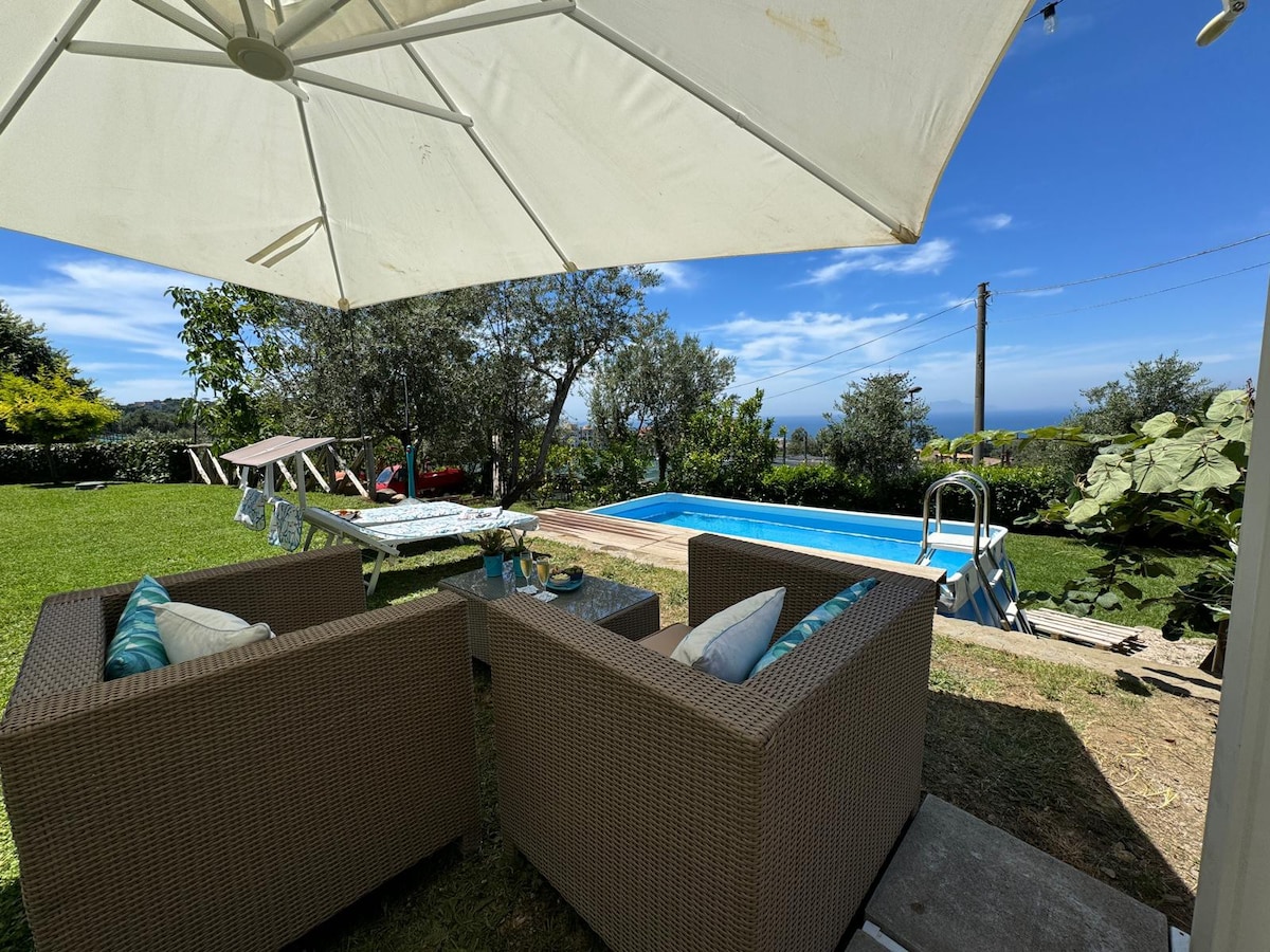 A shaded seating area is presented with comfortable wicker chairs and a small table. A private pool is visible nearby, surrounded by lush greenery, with sun loungers positioned on the pool deck. The sky is bright and blue, adding warmth to the outdoor scene.