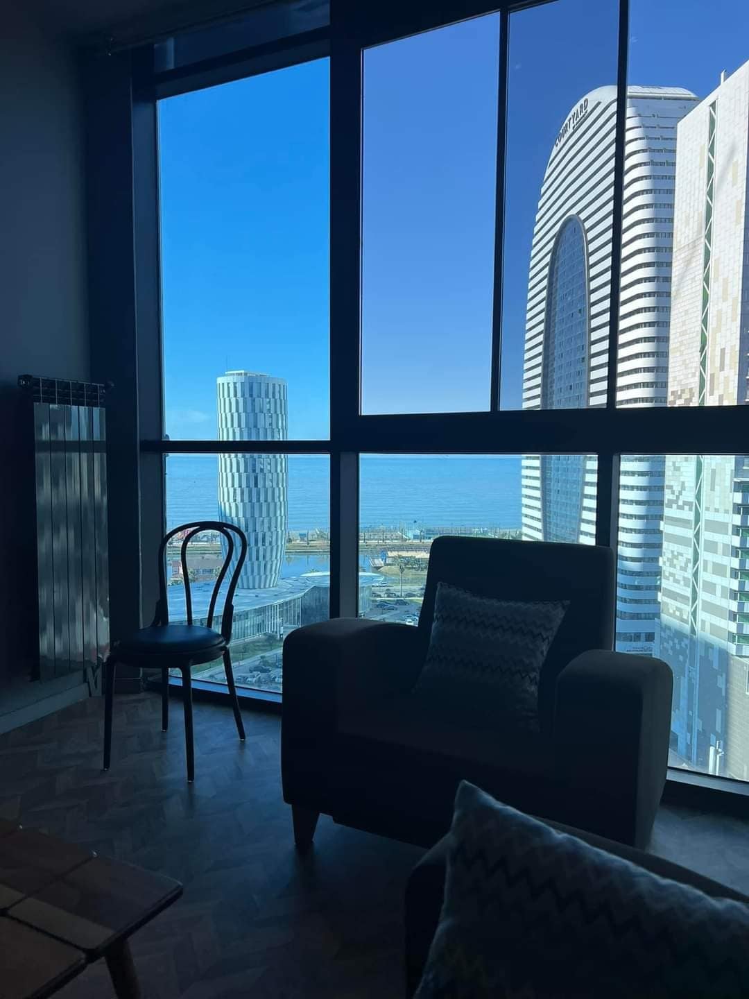 A spacious living area is showcased through large glass windows, offering views of nearby buildings and the sea. A modern armchair and a simple chair are positioned to enjoy the scenery, while natural light illuminates the space.