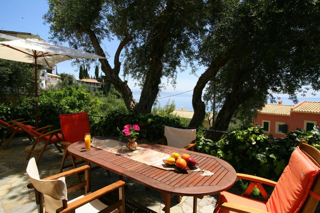 A shaded outdoor dining area features a wooden table set with a variety of fresh fruits and a drink. Surrounding the table are colorful chairs, and the scene is framed by olive trees, with a glimpse of the sea visible in the background.