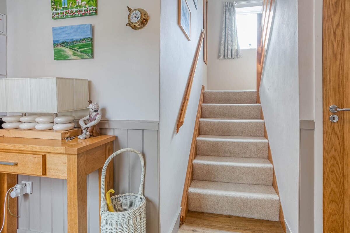 A well-lit hallway features a wooden table with a decorative lamp and a woven basket. A carpeted staircase leads to the upper floor, with a window at the top allowing natural light to fill the space.