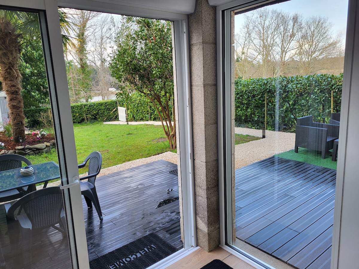 Large glass doors frame a view of a garden featuring a seating area with black chairs and a glass table. Lush greenery surrounds the outdoor space, while a pathway made of gravel leads into the garden, providing a peaceful atmosphere.