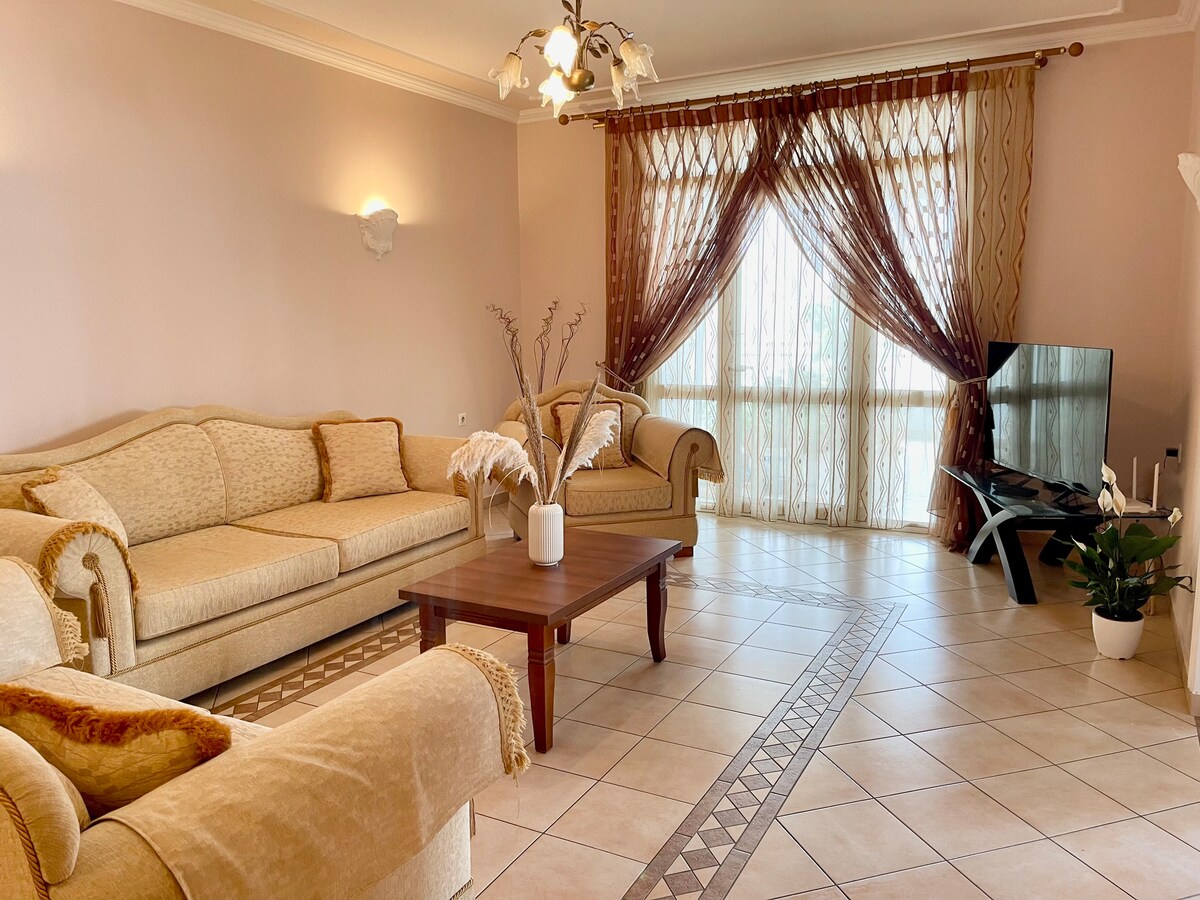 A spacious living area features a light-colored sofa set arranged around a wooden coffee table. Natural light filters in through large, draped windows, creating a bright atmosphere. A modern television is positioned on a sleek stand, and decorative plants add a touch of greenery.