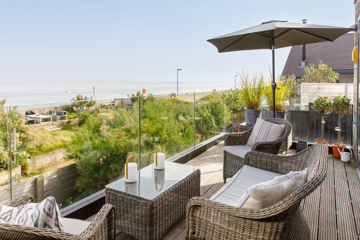 A private terrace offers comfortable seating with woven chairs and a glass-top table. An umbrella provides shade, while coastal views stretch towards the horizon, showcasing beach vegetation and the shimmering sea beyond. Green planters enhance the serene atmosphere.