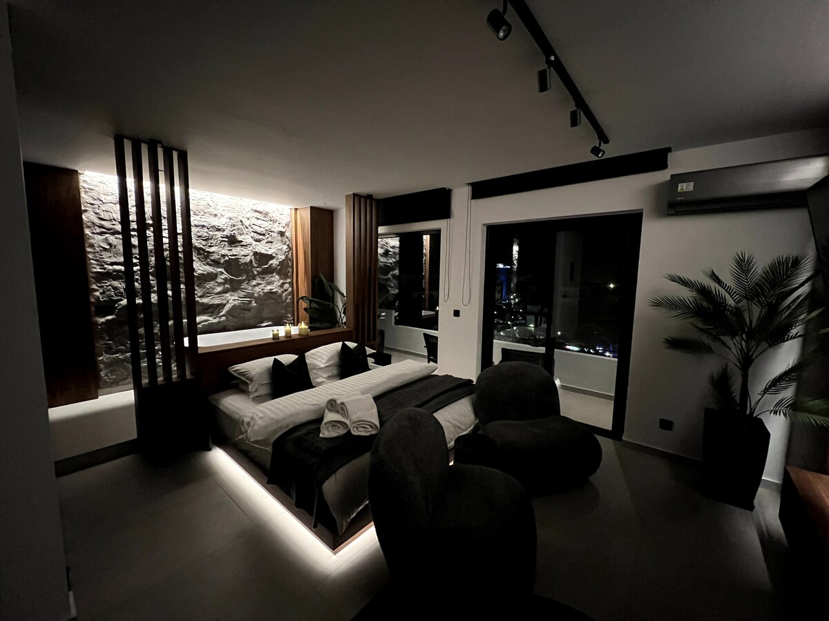 A modern bedroom features a spacious bed, complemented by wooden accents and a striking natural rock wall. Soft ambient lighting creates a serene atmosphere, while large windows offer views of the outside. A plant adds a touch of nature to the space.
